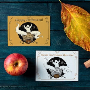 May include: Two Halloween greeting cards with a white ghost in a cauldron with other ghosts around it. The card on the left has a yellow background and says "Happy Halloween!" The card on the right has a white background and says "Ghouls Just Wanna Have Fun".