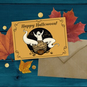 May include: A yellow card with the text "Happy Halloween!" and a drawing of three ghosts in a cauldron with a pumpkin. The card is on a brown envelope and a blue wooden background with fall leaves.