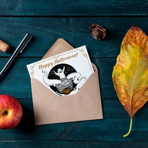 May include: A brown envelope with a white card inside. The card has a black and white illustration of a ghost in a cauldron with the text "Happy Halloween!" in gold lettering.