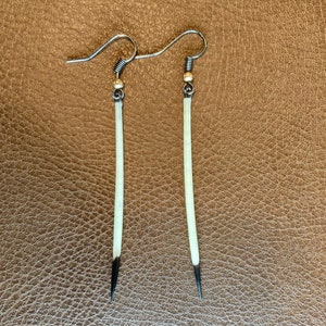 May include: A pair of handmade earrings featuring long, slender, off-white quills with black tips. The quills are suspended from silver-colored ear wires with small gold-colored beads. The earrings are displayed against a brown leather-textured background.