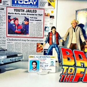 K&ouml;nnte beinhalten: Eine Nahaufnahme einer Back-to-the-Future-Darstellung mit einem silbernen DeLorean-Zeitfahrzeug, zwei Actionfiguren von Marty McFly, einem Zeitungsartikel mit der Schlagzeile "Jugendlicher inhaftiert" und einem kalifornischen F&uuml;hrerschein f&uuml;r Martin McFly. Die Darstellung ist vor einem wei&szlig;en Hintergrund.