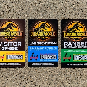 Puede incluir: Tres tarjetas de identificaci&oacute;n rectangulares de Jurassic World. Las tarjetas son marrones con el logotipo de Jurassic World en la parte superior. Las tarjetas dicen "VISITOR", "LAB TECHNICIAN" y "RANGER". Cada tarjeta tiene una franja de color diferente en la parte inferior con texto y un c&oacute;digo de barras.