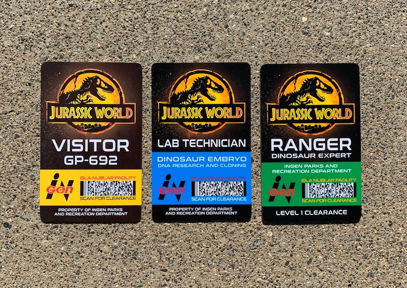 Puede incluir: Tres tarjetas de identificaci&oacute;n rectangulares de Jurassic World. Las tarjetas son marrones con el logotipo de Jurassic World en la parte superior. Las tarjetas dicen "VISITOR", "LAB TECHNICIAN" y "RANGER". Cada tarjeta tiene una franja de color diferente en la parte inferior con texto y un c&oacute;digo de barras.