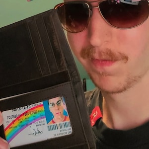 K&ouml;nnte beinhalten: Eine dunkelbraune Lederbrieftasche mit einem transparenten Ausweisfach. Der Ausweis zeigt ein Regenbogenmuster und ein Foto. Die Brieftasche ist ge&ouml;ffnet und zeigt die Karte. Die Person tr&auml;gt eine Sonnenbrille.