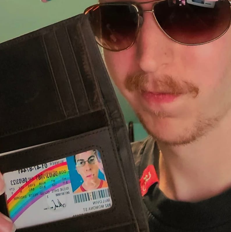K&ouml;nnte beinhalten: Eine dunkelbraune Lederbrieftasche mit einem transparenten Ausweisfach. Der Ausweis zeigt ein Regenbogenmuster und ein Foto. Die Brieftasche ist ge&ouml;ffnet und zeigt die Karte. Die Person tr&auml;gt eine Sonnenbrille.