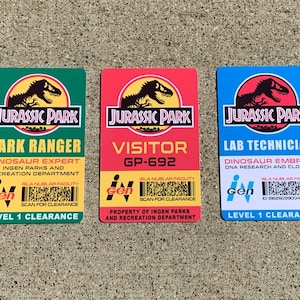 Puede incluir: Tres tarjetas de identificaci&oacute;n de Jurassic Park: verde para Guardabosques, roja para Visitante y azul para T&eacute;cnico de laboratorio. Cada una presenta el logotipo de Jurassic Park y detalles de los roles.