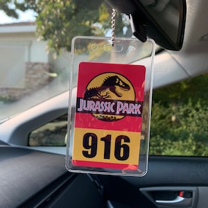 Puede incluir: Un portaetiquetas de coche de pl&aacute;stico transparente con un dise&ntilde;o de Parque Jur&aacute;sico. La etiqueta presenta el logotipo de Parque Jur&aacute;sico con una silueta de T-Rex, las palabras "JURASSIC PARK" y el n&uacute;mero "916" en un esquema de color amarillo y rojo.