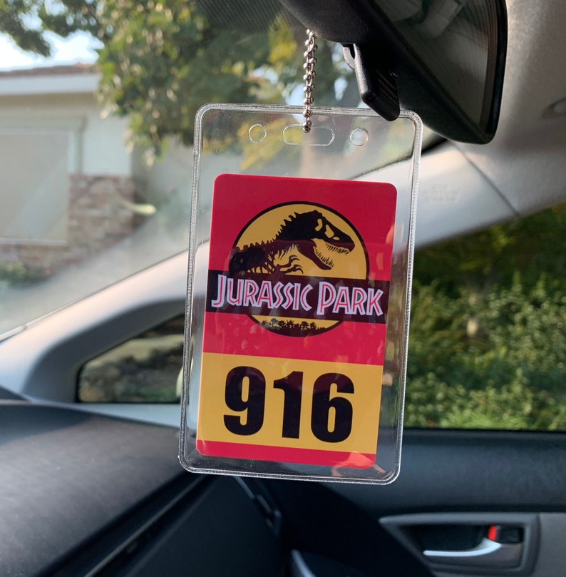Puede incluir: Un portaetiquetas de coche de pl&aacute;stico transparente con un dise&ntilde;o de Parque Jur&aacute;sico. La etiqueta presenta el logotipo de Parque Jur&aacute;sico con una silueta de T-Rex, las palabras "JURASSIC PARK" y el n&uacute;mero "916" en un esquema de color amarillo y rojo.