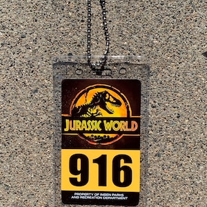Puede incluir: Tarjeta de identificaci&oacute;n de Jurassic World de pl&aacute;stico transparente con una cadena plateada. La tarjeta presenta el logotipo de Jurassic World, el n&uacute;mero 916 y el texto "PROPERTY OF INGEN PARKS AND RECREATION DEPARTMENT."