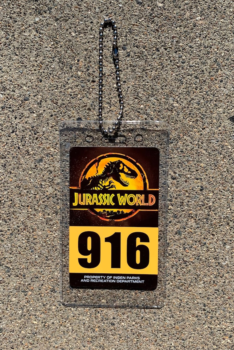 Puede incluir: Tarjeta de identificaci&oacute;n de Jurassic World de pl&aacute;stico transparente con una cadena plateada. La tarjeta presenta el logotipo de Jurassic World, el n&uacute;mero 916 y el texto "PROPERTY OF INGEN PARKS AND RECREATION DEPARTMENT."