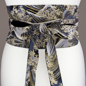 Può includere: Ampia cintura obi con motivo a onde in blu navy, grigio e oro. La cintura è annodata con un fiocco sul davanti, con lunghi lacci pendenti. Il tessuto ha una lucentezza sottile e il design è dettagliato ed elegante. Un accessorio in stile giapponese.
