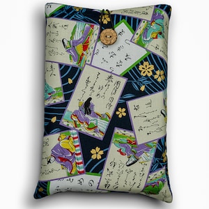 May include: A rectangular book sleeve with a Japanese-inspired design. The fabric features illustrations of figures in traditional attire, calligraphy, and floral patterns in shades of purple, green, and gold against a navy blue background. It has a wooden button closure.
