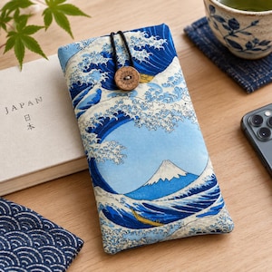 May include: A rectangular fabric pouch featuring a blue and white print of a large ocean wave and a mountain. The pouch is secured with a black elastic cord looped over a round, light brown wooden button with a floral pattern engraved on it.