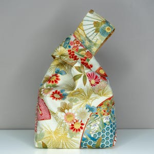 May include: A Japanese-style knot bag made of patterned fabric. The bag features a cream background with colorful floral and fan motifs in red, blue, and gold. The bag is folded at the top to create a handle.
