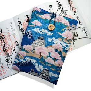 May include: A rectangular fabric sleeve featuring a blue background with a print of Japanese castles, cherry blossom trees, and a mountain. The sleeve is secured with a black elastic cord and a round, light-coloured wooden button closure.