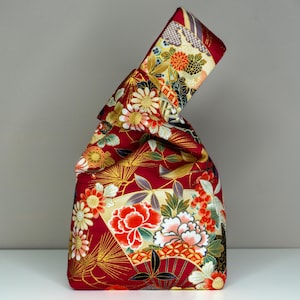 May include: A red fabric bag with a Japanese floral pattern. The bag features a repeating design of flowers, fans, and gold accents. The bag is folded at the top, creating a unique shape. The bag is made of a sturdy material.