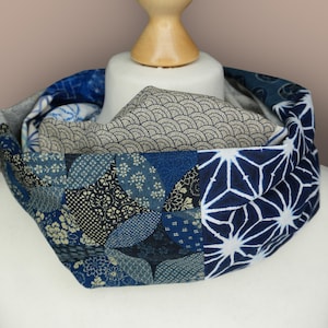Puede incluir: Un pañuelo infinito de patchwork en tonos azules, grises y blancos. El pañuelo presenta varios patrones de inspiración japonesa, incluyendo diseños florales, geométricos y de olas. El pañuelo se muestra en una cabeza de maniquí blanca.
