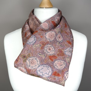 Puede incluir: Bufanda estampada en tonos malva, rosa y azul, colocada sobre un maniquí. La bufanda presenta un diseño floral repetido con flores y hojas estilizadas. La tela tiene una sutil apariencia texturizada, agregando profundidad al diseño.