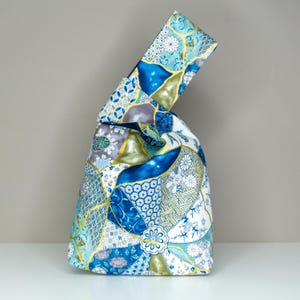 Könnte beinhalten: Eine japanische Knotentasche im Patchwork-Design. Die Tasche weist eine Vielzahl von blauen, weißen und goldenen Blumen- und geometrischen Mustern auf. Die Tasche ist aus Stoff gefertigt und hat einen einzigartigen Knotenverschluss.
