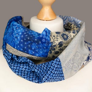 Puede incluir: Bufanda infinita con estampado azul y blanco. La bufanda presenta varios diseños geométricos y florales en tonos azules, blancos y beige. La bufanda está hecha de una tela suave y se muestra en una cabeza de maniquí.