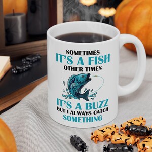 May include: White ceramic mug with a black liquid inside. The mug features the text "SOMETIMES IT'S A FISH OTHER TIMES IT'S A BUZZ BUT I ALWAYS CATCH SOMETHING" and a blue fish graphic. The mug is on a gray surface with Halloween-themed candies.