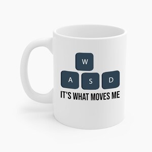 Pode incluir: Caneca de cerâmica branca com uma alça preta. A caneca apresenta um design de quatro teclas de teclado azul escuro com as letras "W", "A", "S" e "D". Abaixo das teclas está o texto "IT'S WHAT MOVES ME" em preto.
