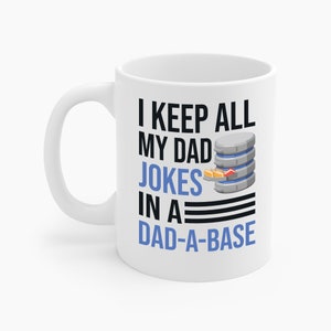 Op de afbeelding: Witte keramische mok met de tekst "I KEEP ALL MY DAD JOKES IN A DAD-A-BASE" in zwart en blauw. Een afbeelding van een databaseserver staat aan de rechterkant van de mok.
