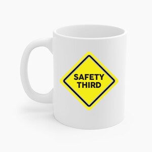 Puede incluir: Taza de cerámica blanca con un diseño en forma de diamante amarillo. El diseño tiene un borde negro y las palabras "SAFETY THIRD" en letras negras en negrita. La taza tiene un asa curvada.