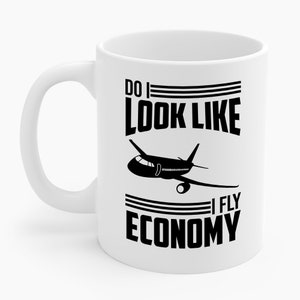 Pode incluir: Caneca de cerâmica branca com um gráfico de avião preto e o texto "DO I LOOK LIKE I FLY ECONOMY". A caneca tem uma alça curva e uma forma cilíndrica. O texto e o gráfico estão centrados na caneca.