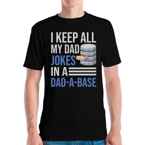 Puede incluir: Camiseta negra con el texto "I KEEP ALL MY DAD JOKES IN A DAD-A-BASE" en blanco y azul. El gráfico incluye un icono de servidor de base de datos con libros. Una persona lleva la camiseta.