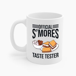 May include: White ceramic mug with the text "OFFICIAL S'MORES TASTE TESTER" in black. The mug features a graphic of marshmallows, chocolate, and s'mores. The mug has a curved handle and is designed for hot beverages.