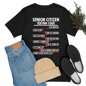 Puede incluir: Camiseta negra con el texto "SENIOR CITIZEN TEXTING CODE" en blanco. La camiseta presenta una lista de acrónimos de mensajes de texto y sus significados. La camiseta se combina con vaqueros azules, un gorro beige y zapatos negros.