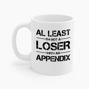 May include: White ceramic mug with a black text that reads "AL LEAST I'M NOT A LOSER WITH AN APPENDIX". The mug has a curved handle and a cylindrical shape. The text is in a bold, distressed font.