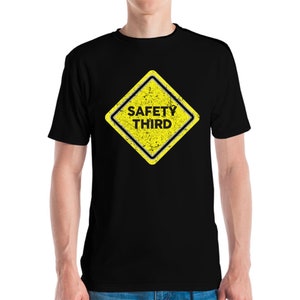 Puede incluir: Camiseta negra con un letrero amarillo en forma de diamante con la frase "SAFETY THIRD" en negro. El letrero tiene un efecto desgastado. La camiseta es de cuello redondo.