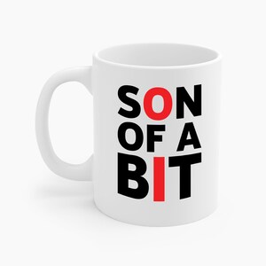 May include: White ceramic mug with the text "SON OF A BIT" in black and red block letters. The mug has a curved handle and a glossy finish. The mug is designed for hot beverages.