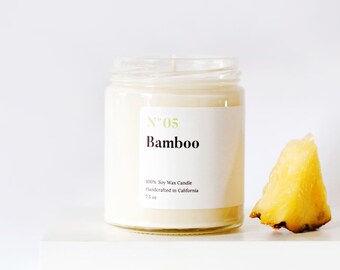 May include: A clear glass jar candle with a white label that reads "N°05 Bamboo". The candle is filled with a creamy wax and has a single wick. A piece of pineapple sits next to the candle. The candle is 7.5 oz.