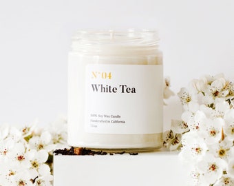 Peut inclure: Une bougie en cire de soja blanche dans un pot en verre transparent avec une étiquette blanche sur laquelle est inscrit "N°04 White Tea 100% Soy Wax Candle Handcrafted in California 7.5 oz".