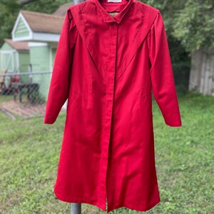 May include: A long red coat with a button-up front and a collar. The coat has a slightly flared hem and long sleeves.