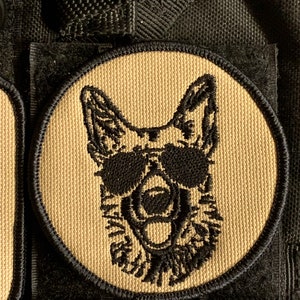 May include: A tan and black embroidered patch of a German Shepherd dog wearing sunglasses. The dog is looking forward with its mouth open and tongue out. The patch is circular and has a black border.
