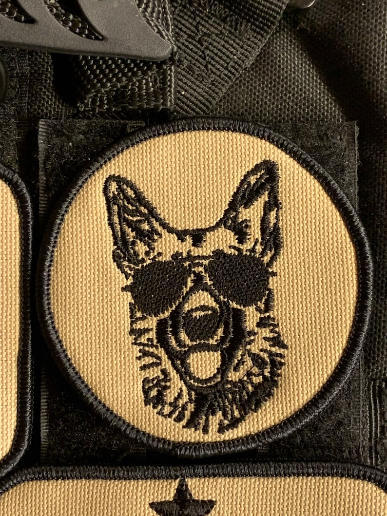 May include: A tan and black embroidered patch of a German Shepherd dog wearing sunglasses. The dog is looking forward with its mouth open and tongue out. The patch is circular and has a black border.