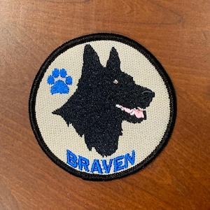 May include: A round, beige embroidered patch with a black German Shepherd silhouette, a blue paw print, and the word "BRAVEN" in blue lettering.