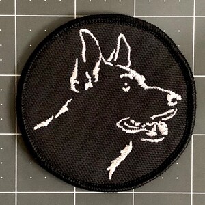 May include: A black and white embroidered patch of a German Shepherd dog's head. The dog is looking to the right and has its mouth slightly open.