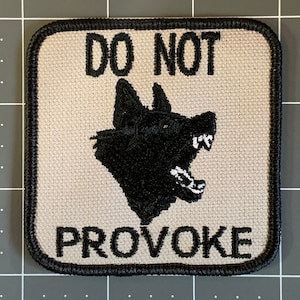May include: A white embroidered patch with black stitching. The patch features a black wolf's head with a menacing expression and the text "DO NOT PROVOKE" in black.