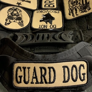 May include: Black tactical dog harness with a tan patch that reads "GUARD DOG". The harness has several other tan patches with text and images, including "DO NOT PROVOKE", "BRAVE", "COME AND TAKE", and a wolf silhouette.