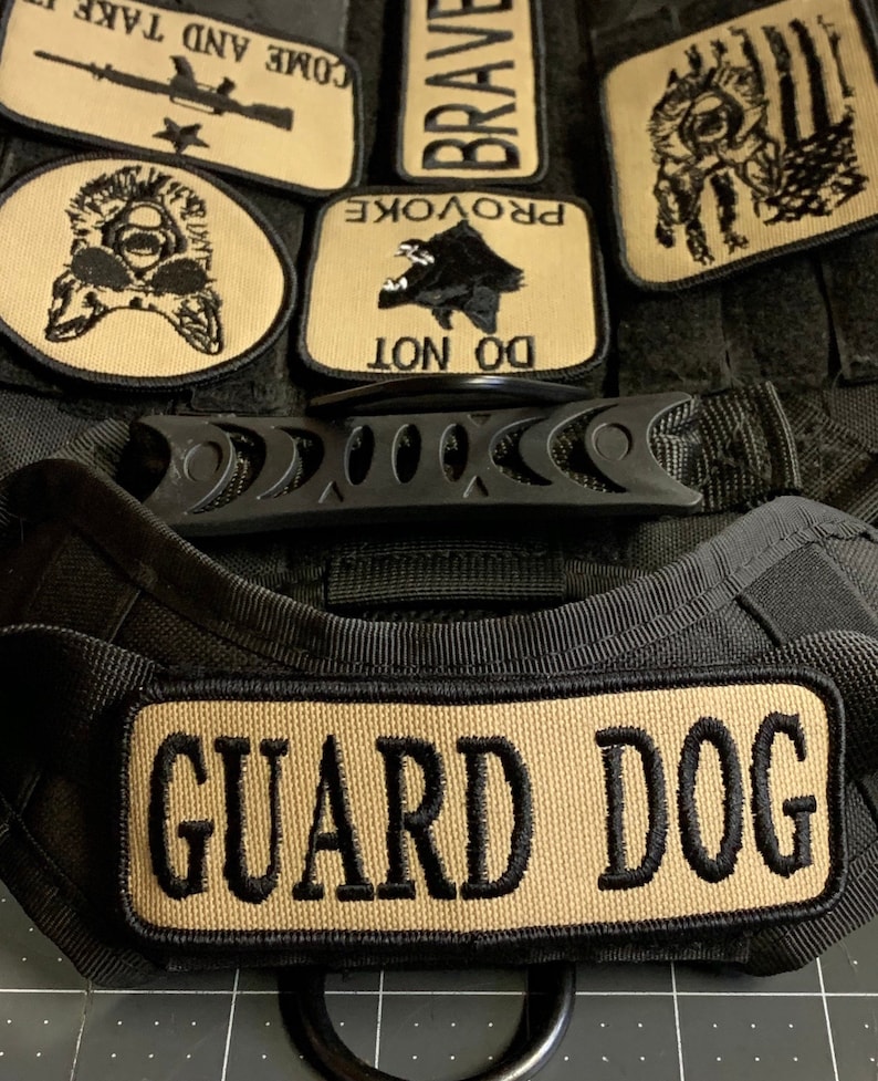 May include: Black tactical dog harness with a tan patch that reads "GUARD DOG". The harness has several other tan patches with text and images, including "DO NOT PROVOKE", "BRAVE", "COME AND TAKE", and a wolf silhouette.