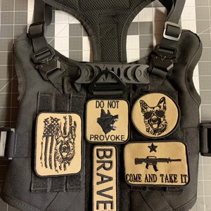 May include: Black tactical dog harness with multiple patches. The patches include a dog silhouette with the text "Do Not Provoke", a dog with sunglasses, a dog with an American flag, a patch with the text "Braven", and a patch with a rifle and the text "Come and Take It".