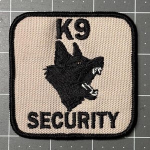 May include: A square patch with a black border features the text "K9 SECURITY" in dark gray. A black silhouette of a snarling dog is centered above the word "SECURITY". The background is a light tan color.