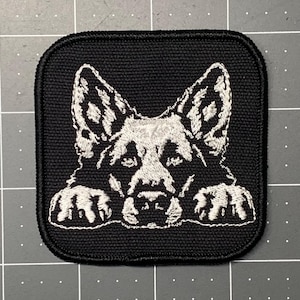May include: A black embroidered patch with a white German Shepherd dog peeking over the top. The dog's paws are visible on either side of the patch.