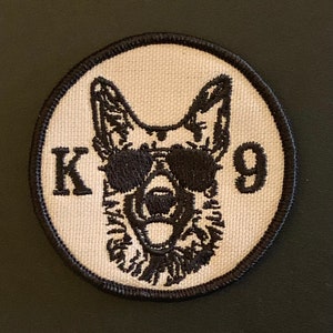 May include: A white embroidered patch with a black German Shepherd wearing sunglasses. The patch says "K9" in black lettering.