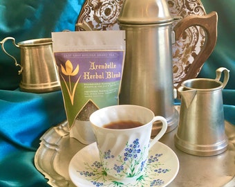 May include: A silver tea pot, a silver creamer, and a silver cup with a floral design on a silver tray. A tea bag package with the text "Lady Gray Kitchen Brand Tea Arendelle Herbal Blend" is on the tray.